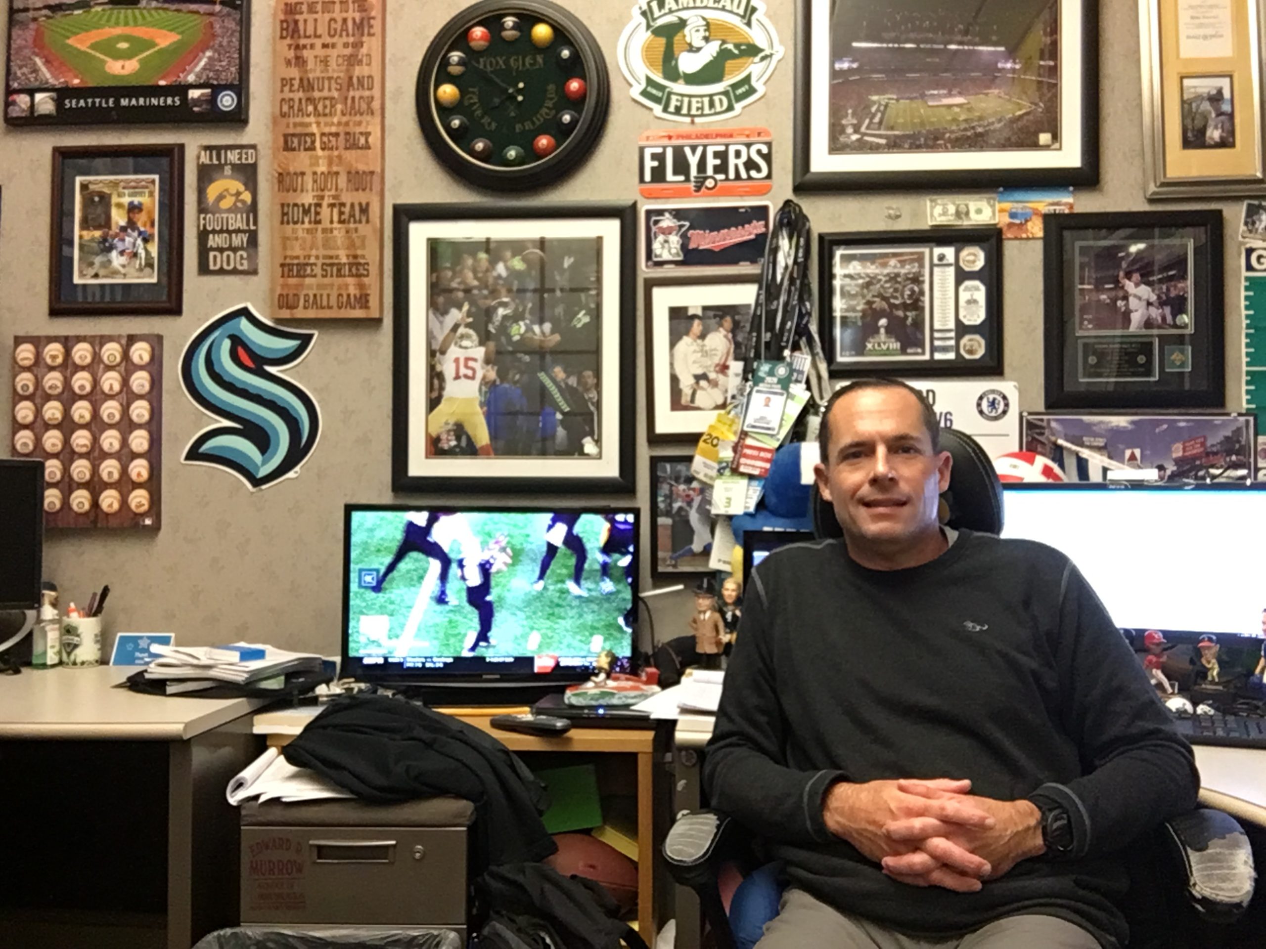 An image of a male seated in front of the camera with a desk set-up behind him surrounded by Seattle sports memorabilia.